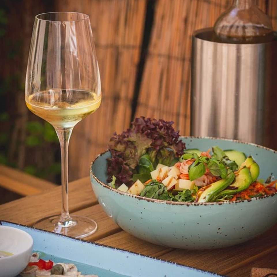 A glass of white wine next to a fresh Italian salad with avocado, shrimp, and cheese.